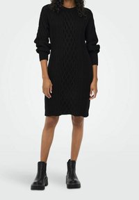 Robe-pull noire tricotée avec motif torsadé, col rond et manches longues bouffantes, associée à des bottines noires épaisses.