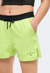 Femme portant un short de sport Puma HYROX vert néon avec une ceinture noire et les mains dans les poches, cadrée à mi-corps.