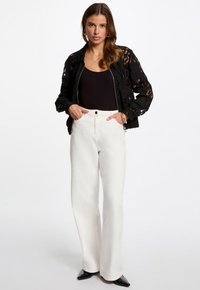 Veste bomber en dentelle noire sur un haut noir, associée à un pantalon large taille haute blanc et à des chaussures noires à bout pointu.