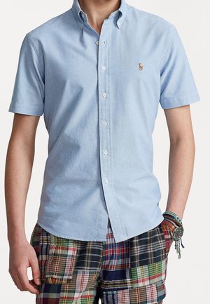 Mann trägt hellblaues, kurzärmeliges Hemd mit besticktem Logo, mehrfarbige Patchwork-Shorts und Armbänder mit Armbanduhr.