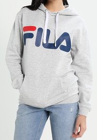 Sudadera gris con un gran logotipo de FILA en azul marino y rojo, capucha con cordón, bolsillos frontales y puños acanalados. Combinada con jeans claros de mezclilla desgastados.