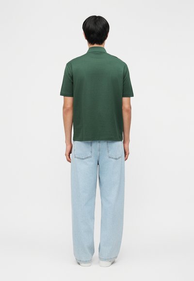 Homme de dos portant un polo à manches courtes vert foncé, un jean large bleu clair et des baskets blanches sur un fond uni.