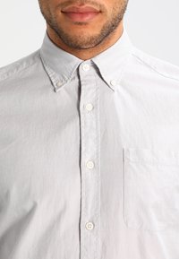 Koszula na krótki rękaw, jasnoszara, zapinana na guziki, z kołnierzykiem, białymi guzikami oraz kieszenią na lewej piersi. Wykonana z teksturowanej tkaniny.