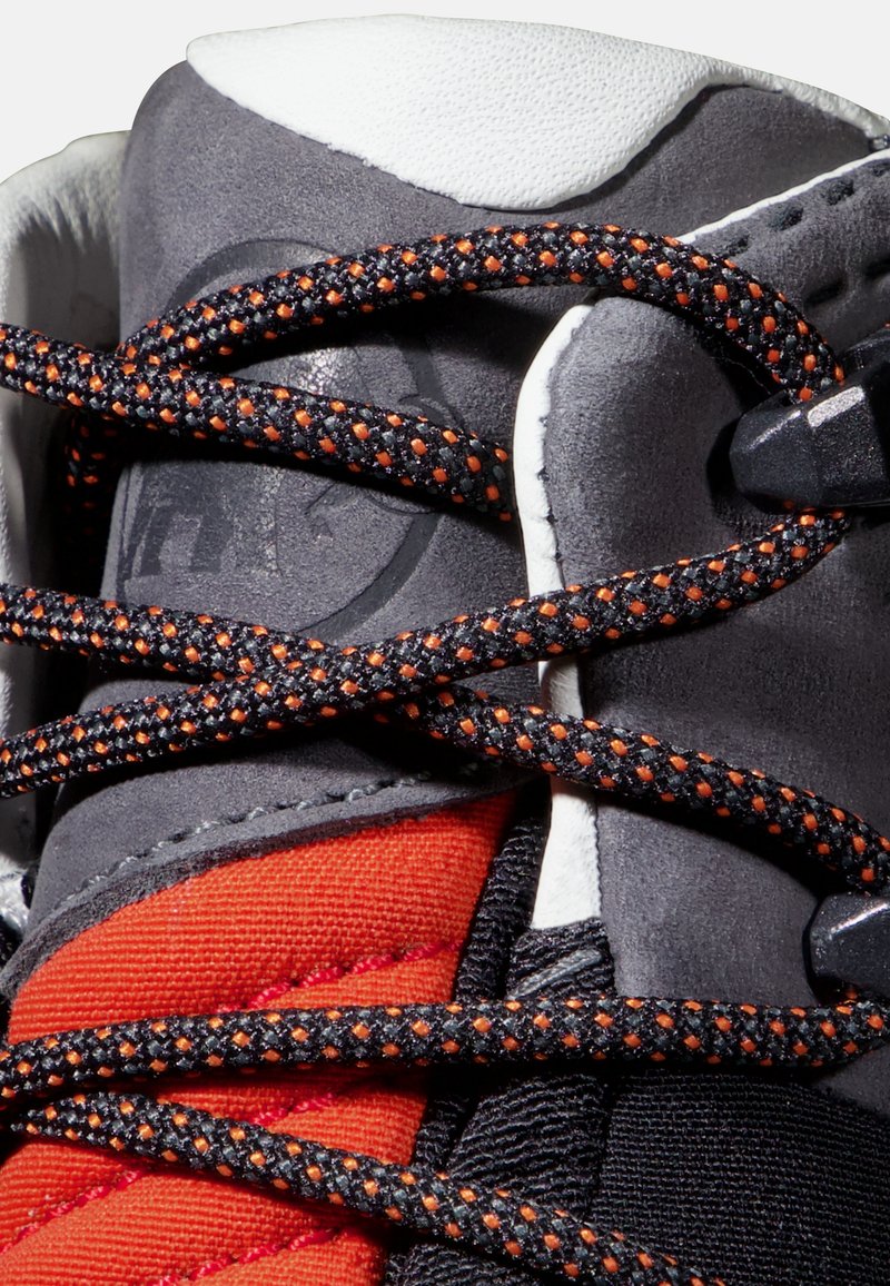 Scarpa grigia e nera con sezioni in tessuto rosso, caratterizzata da lacci neri con macro fiori arancioni. Materiali testurizzati e dettagli di design robusti visibili.