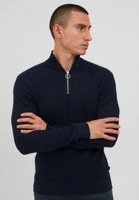 Vīrietis ar īsiem matiem, ģērbies tumš zilā struktūrēta zip-up džemperī ar gredzenu zipsa vilkšanai, ar rokām krustām, skatās pa labi uz vienkāršas fona.