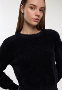 Młoda kobieta z prostymi brązowymi włosami, ubrana w czarny puszysty sweter, pozująca z rękami na biodrach na jednolitym tle.