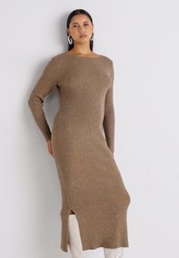 Robe en tricot à manches longues dans une texture côtelée marron clair, avec un col bateau et une fente latérale, accompagnée de pantalons ajustés de couleur crème.