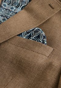 Blazer texturé marron avec un mouchoir de poche à motif floral bleu et noir glissé dans la poche poitrine.