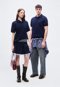 Γυναίκα και άνδρας που φορούν ναυτικές πόλο μπλούζες· η γυναίκα με πλισέ φούστα και μπότες κρατάει μια κόκκινη τσάντα, ο άνδρας με καρό πουκάμισο δεμένο γύρω από τη μέση και τζιν παντελόνι.