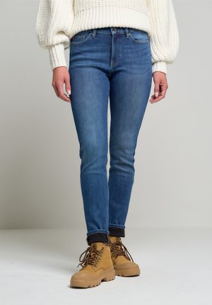 Nainen, joka pukeutunut istuviin sinisiin farkkuihin, beigen värisiin nyörikengännauhoihin ja valkoiseen neulepuseroon, seisoo yksinkertaisen vaaleanharmaan taustan edessä.