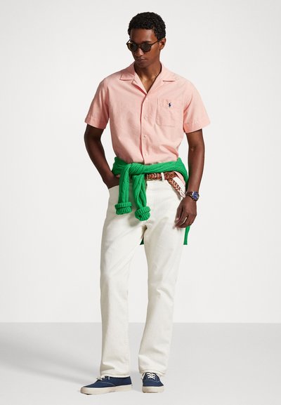 Ροζ κοντομάνικο πουκάμισο με τσέπη στο στήθος, λευκά παντελόνια, πράσινο πουλόβερ δεμένο στη μέση και μπλε αθλητικά παπούτσια. Τα γυαλιά ηλίου συμπληρώνουν την εμφάνιση.