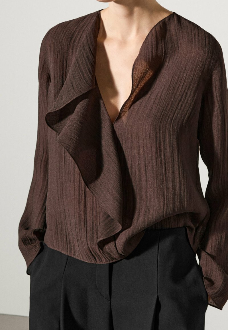 Blouse marron à manches longues en maille côtelée avec un décolleté en V profond et un détail drapé à l'avant, associée à un pantalon noir taille haute.