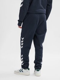 Man wearing navy blue track pants with white chevron stripes on sides and white sneakers with dark green accents, standing against plain background.