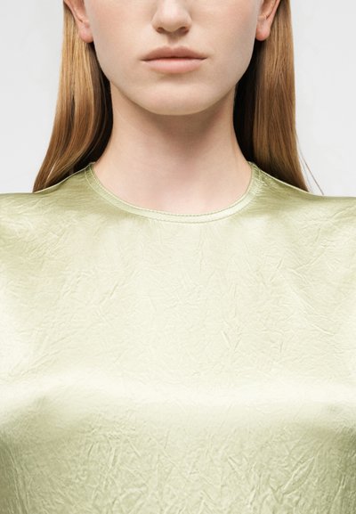 Νεαρή γυναίκα με ίσια ξανθά μαλλιά που φοράει ένα ανοιχτό πράσινο, υφασμάτινο, γυαλιστερό τοπ, εμφανιζόμενη από τον λαιμό μέχρι το στήθος με φόντο ένα ανοιχτόχρωμο φόντο.