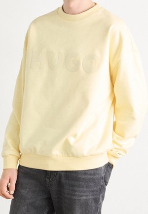 Mand iført lysegul sweatshirt med præget "HUGO"-tekst og mørke jeans, stående med den ene hånd i lommen mod en hvid baggrund.