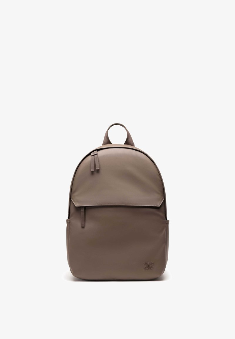 Brown backpack made of smooth, durable material. Features a rounded shape, front flap, zipper, and a top carrying handle. Minimalist design.