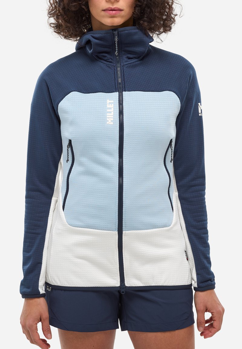 Woman wearing a blue, light blue, and white zip-up fleece jacket with pockets and navy shorts, standing against a white background.