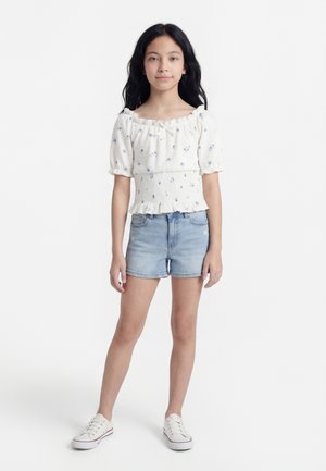 Mädchen mit langen schwarzen Haaren, trägt eine weiße Bluse mit Puffärmeln und Blumenmuster, hellblaue Denim-Shorts und weiße Turnschuhe, steht vor weißem Hintergrund