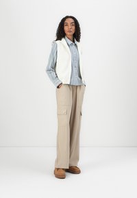 Gilet en tricot côtelé blanc, chemise à boutons rayée bleue, pantalon cargo beige et chaussures à plateforme brunes. Comprend des poches et une coupe surdimensionnée.