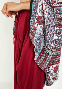 Manos entrelazadas sobre pantalones rojos fluidos, con una bufanda estampada que presenta diseños florales y geométricos en rojo, azul y negro, descansando a su lado.