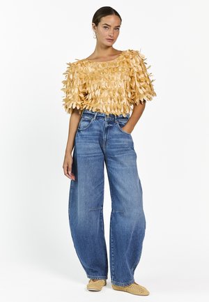 Vrouw die een beige gestructureerde top met korte mouwen draagt, wijde blauwe jeans, beige gevlochten schoenen en parel druppel oorbellen, staand met één hand in de zak.