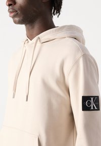 Felpa con cappuccio beige realizzata in tessuto morbido, con tasca a marsupio, cappuccio con lacci e una cucitura nera con logo sulla manica sinistra.