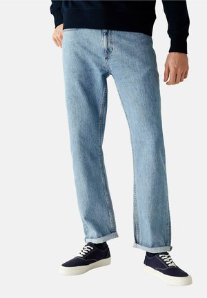 Man draagt lichtblauwe rechte spijkerbroek met omslag en zwarte canvas sneakers met witte zolen en veters, staand tegen een witte achtergrond.