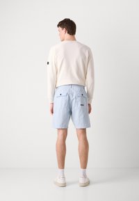 Calções azul claro com dois bolsos traseiros, combinados com um sweatshirt bege claro. O calçado é branco, com uma marca visível de lado.