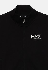 Črna jopica z zadrgo iz flisa z visokim ovratnikom, na levi strani prsnega predela pa je izstopajoči beli logotip EA7 Emporio Armani. Gladka tekstura.