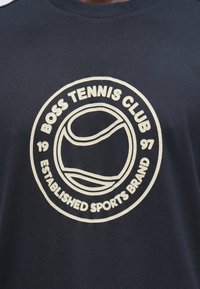 Tumesinine dressipluus valge ümmarguse logoga, millel on tennispall ja tekst "BOSS TENNIS CLUB 1997 ASUTATUD SPORDIBRÄND."