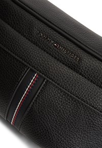 Μαύρη δερμάτινη τσάντα με υφή, οριζόντια ρίγα με κόκκινες, λευκές και μπλε κλωστές, και λογότυπο σε ανάγλυφο στο μπροστινό μέρος.