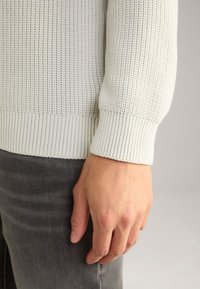 Maglione grigio chiaro lavorato a maglia con motivo testurizzato e polsini a coste, abbinato a jeans grigio scuro. Focus sui dettagli del tessuto e della vestibilità.