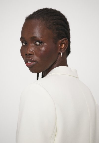 Kvinde med flettet hår iført hvid blazer, kigger over skulderen til siden, med sølv ørering i hoop-stil, mod en ensfarvet baggrund.