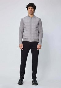 Veste bomber grise matelassée avec des manches côtelées, fermeture éclair avant et deux poches latérales, associée à un pantalon noir et des chaussures foncées.