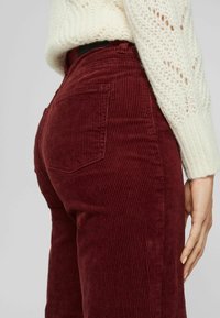 Gros plan sur une personne portant un pantalon en velours côtelé bordeaux taille haute et un pull en tricot blanc, main reposant sur le côté.