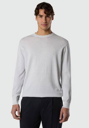 Giovane uomo con capelli castani corti che indossa un maglione bianco a maniche lunghe semplice e pantaloni neri, rivolto in avanti su uno sfondo neutro.