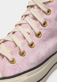 Κοντινό πλάνο ροζ παπουτσιού τύπου ψηλής μπότας με κρέμ κορδόνια περασμένα μέσα από χρυσά ειδώλια και μπεζ λαστιχένια σόλα με μαύρο περίγραμμα.