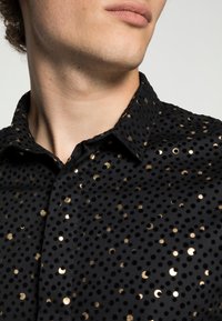 Camisa negra de tela texturizada con prominentes lunares dorados, que cuenta con un cuello clásico y cierre frontal con botones.