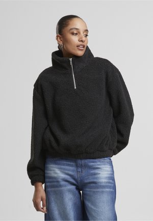 Nő fekete polár pulóvert visel, amelyen fél cipzár van, és kék farmert, egy egyszínű világosszürke háttér előtt áll.