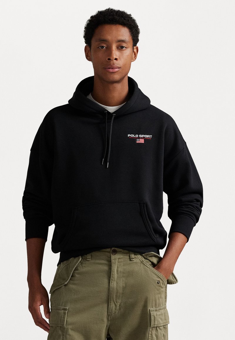 Hanorac negru cu logo-ul "POLO SPORT" și accent cu steagul american. Dispune de un glugă cu șnururi și buzunar tip kangaroo, asortat cu pantaloni cargo de culoare măslinie.
