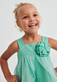 Abito turchese con corpetto aderente, spalline larghe e sovrapposizione di tulle. Presenta un grande fiore decorativo realizzato con lo stesso materiale.