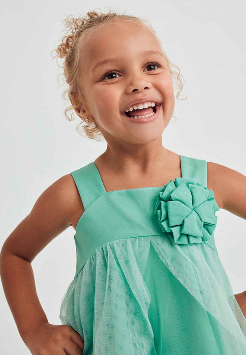 Abito turchese con corpetto aderente, spalline larghe e sovrapposizione di tulle. Presenta un grande fiore decorativo realizzato con lo stesso materiale.