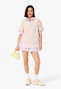 Helles rosa Poloshirt mit kurzen Ärmeln und Tasche, über einem pink-weiß gestreiften Button-up-Shirt getragen, kombiniert mit rosa Shorts und einer gelben Handtasche.