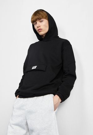Felpa nera con cappuccio in tessuto morbido, con tasca frontale e patch con logo bianco. Abbinata a pantaloni della tuta grigi chiari, a vestibilità comoda.