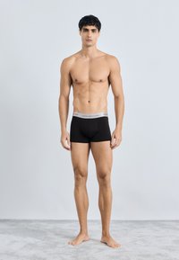 Boxer briefs de algodón negro con una cinturilla gris que presenta el logo de "TOMMY HILFIGER". El modelo está descalzo sobre una alfombra gris.