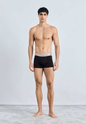 Črne bombažne bokserice z sivo elastiko, na kateri je napis "TOMMY HILFIGER". Model stoji bos na sivi preprogi.