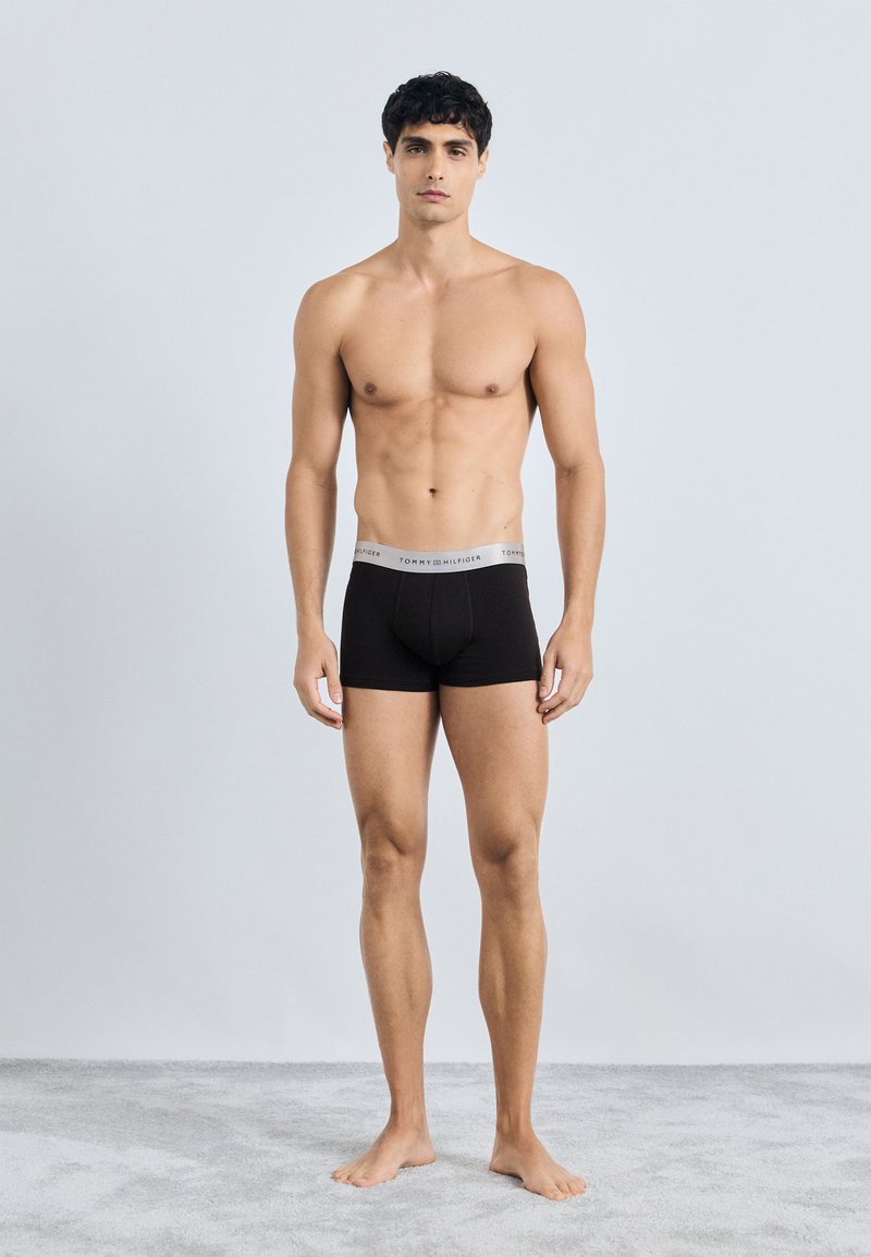 Boxer briefs de algodón negro con una cinturilla gris que presenta el logo de "TOMMY HILFIGER". El modelo está descalzo sobre una alfombra gris.