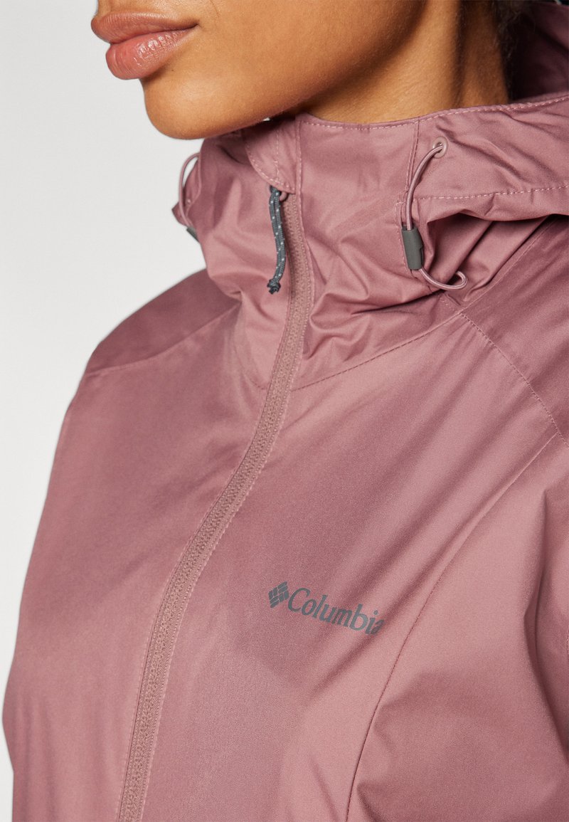 Veste Columbia légère, rose, avec un col haut, fermeture éclair et cordon de réglage. Présente des détails de logo et une texture de tissu lisse.