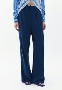 Marineblauwe wijde broek gemaakt van gladde stof, met voorplooien en een hoge taille. De outfit wordt aangevuld met blauwe sandalen met open tenen.