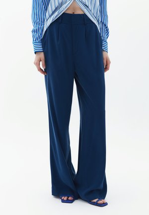 Pantalons évasés bleu marine en tissu lisse, avec des plis sur le devant et une taille haute. La tenue est complétée par des sandales ouvertes bleu. 